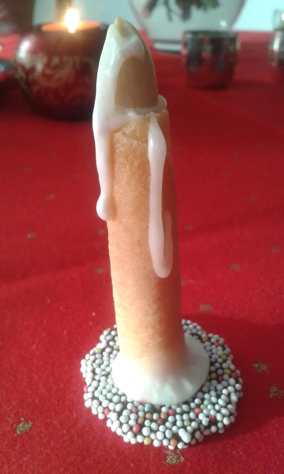 A close-up of a finger-shaped cake or biscuit standing upright on a red tablecloth, decorated with white icing dripping down…