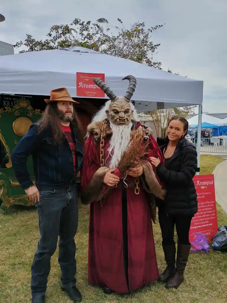 Three people pose outdoors under a white tent: a man in a hat and denim jacket, a Krampus figure in a red robe with horns…
