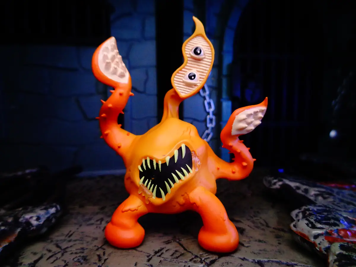 An orange plastic toy monster with a wide mouth full of yellow teeth, two stalked eyes, and clawed limbs stands on a stone…