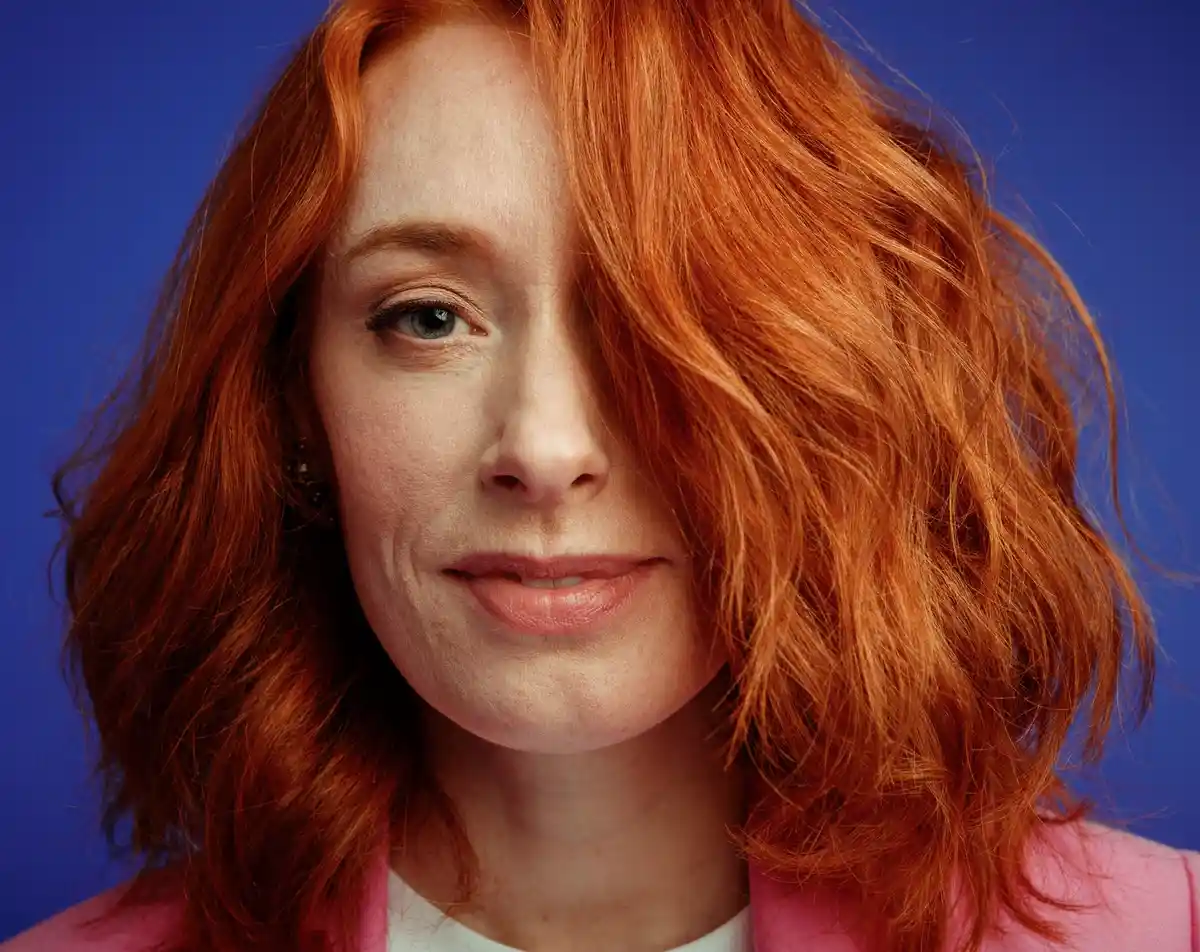 A close-up portrait of a woman with vibrant red hair and blue eyes, wearing a pink jacket over a white shirt, against a…