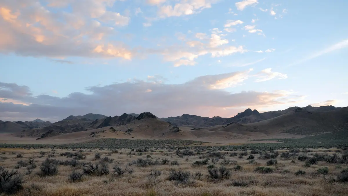 Golden light casts long shadows across the rust-hued dunes as mountains rise like giants from the desert floor, their peaks…