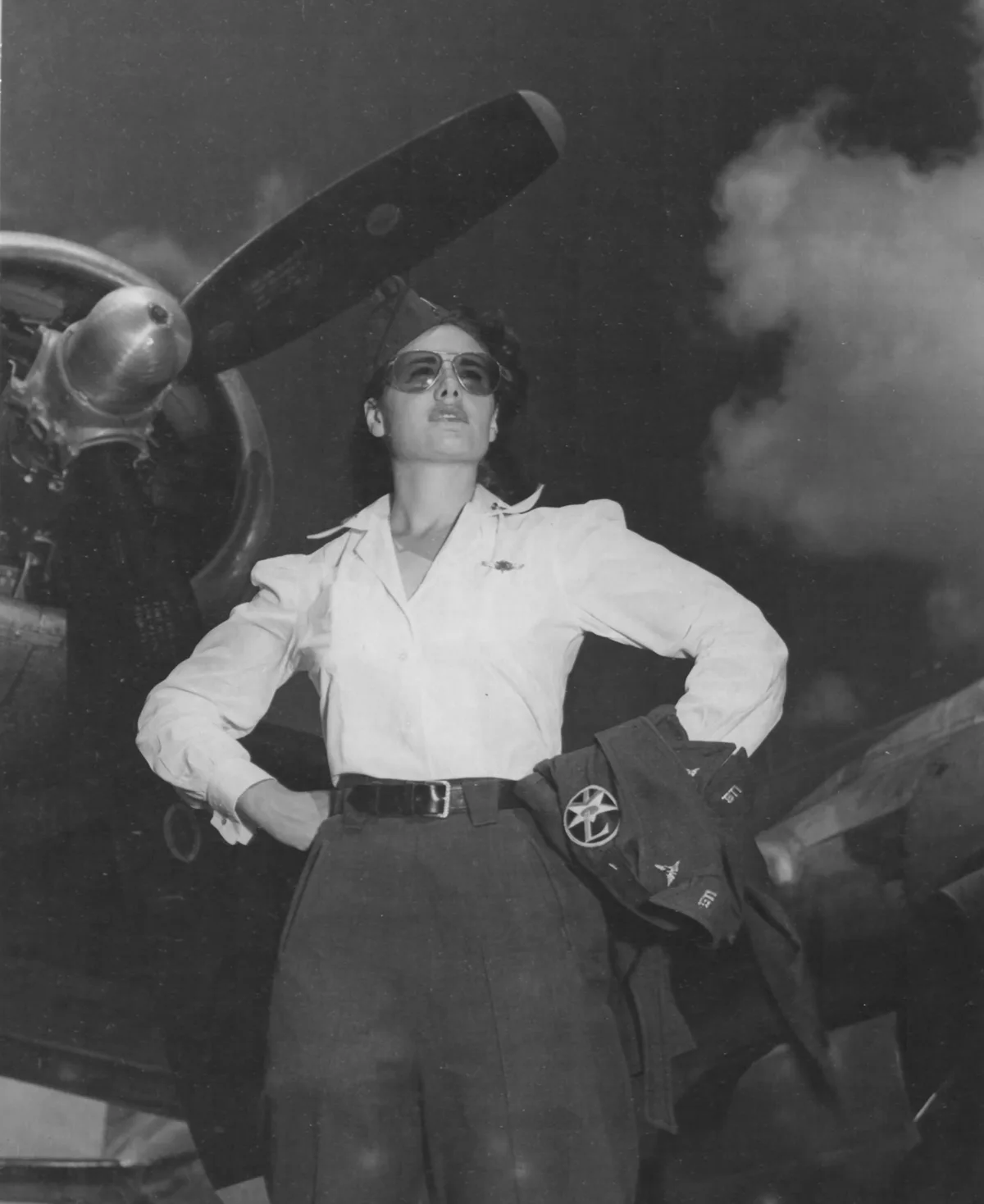 A black-and-white historical photo shows a confident female military pilot in uniform, standing with hands on hips beside a…
