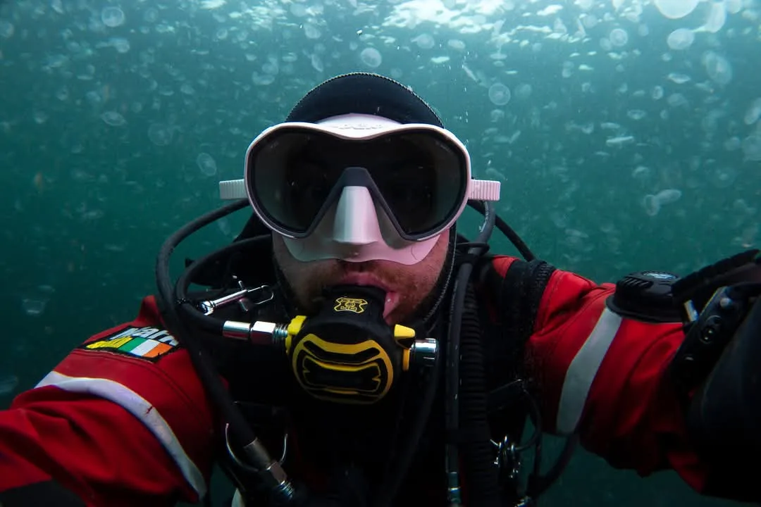 A scuba diver in a red wetsuit and large white mask takes a selfie underwater, surrounded by numerous translucent jellyfish…