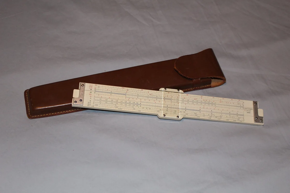 A vintage slide rule with multiple logarithmic scales lies partially extended on a light gray fabric surface, next to its…
