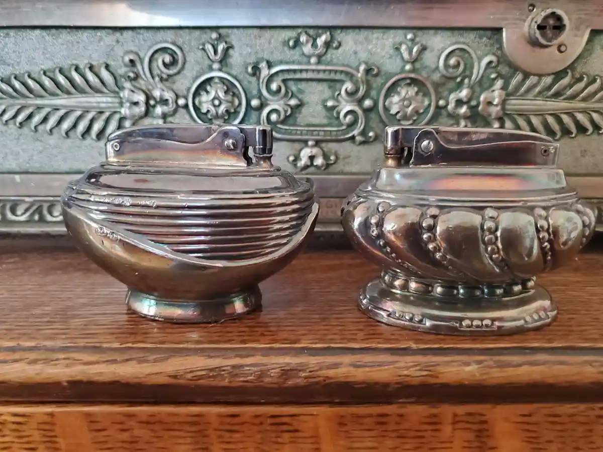 Two ornate, silver-colored salt and pepper shakers sit side-by-side on a wooden surface, each with a hinged lid and…
