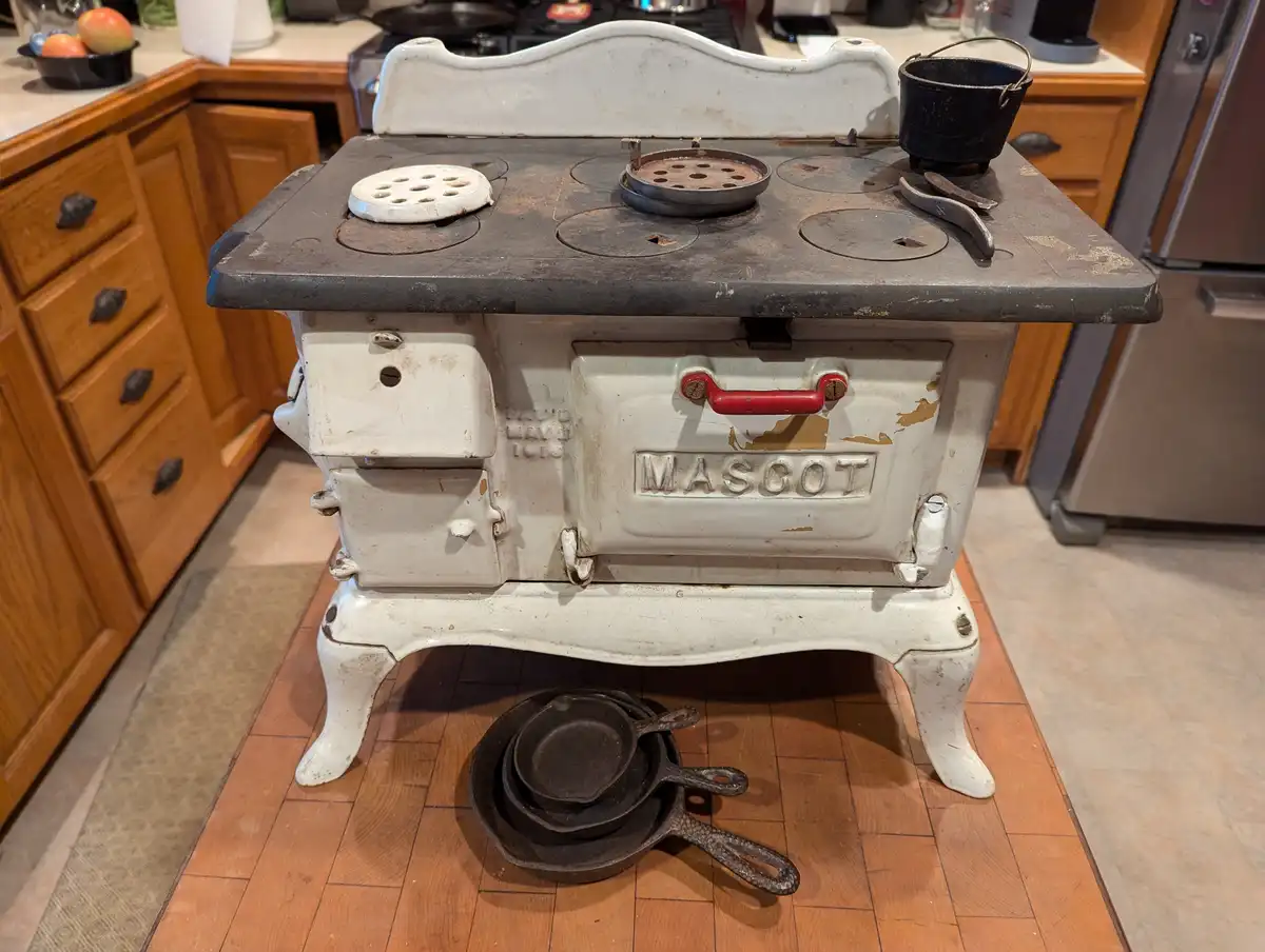 A vintage white cast iron 'MASCOT' stove with a red handle sits in a modern kitchen, showing signs of age and use. On its…