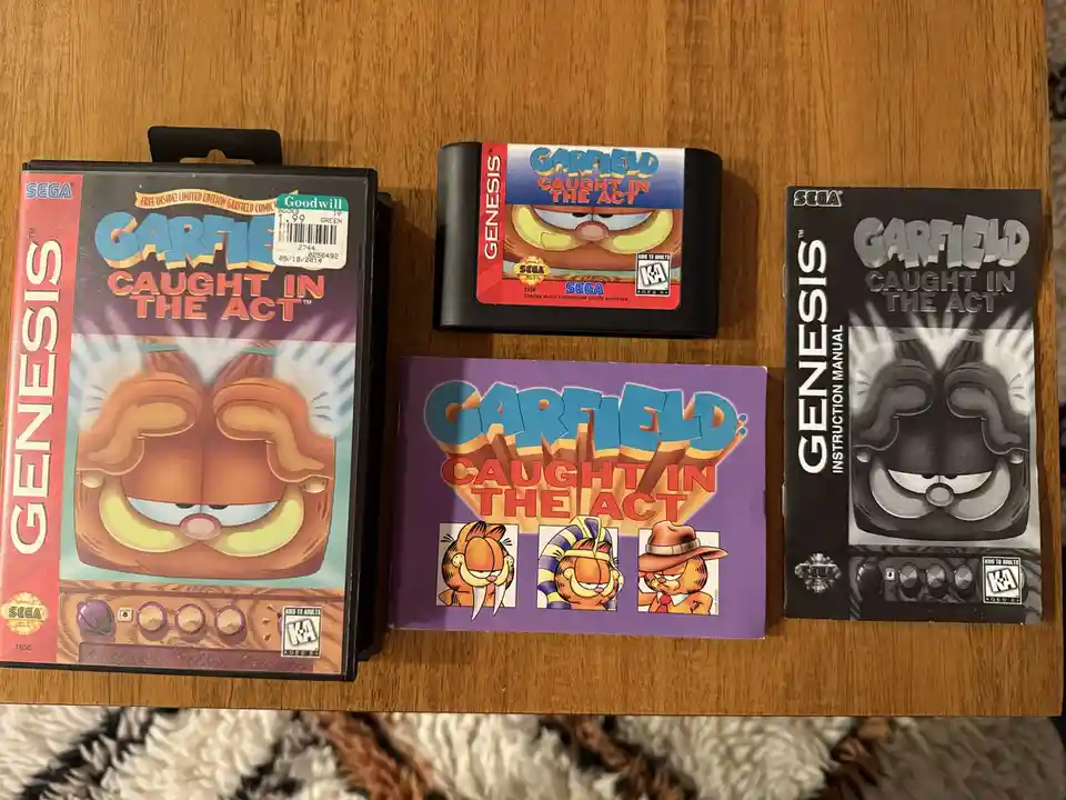 A photo displays the complete Sega Genesis game set for 'Garfield: Caught in the Act', including the game box, cartridge…