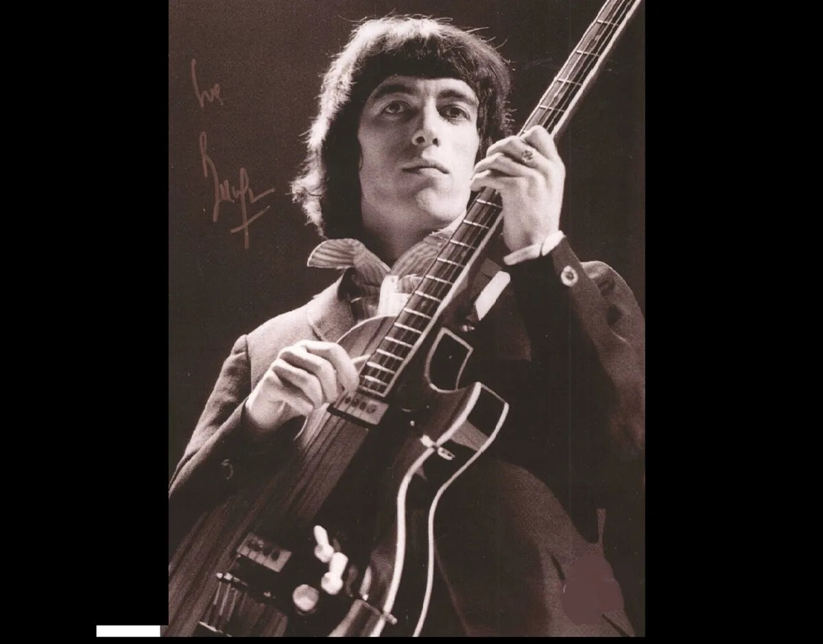A sepia-toned photograph shows a man with shaggy hair holding an electric guitar, wearing a suit and looking upward. His…