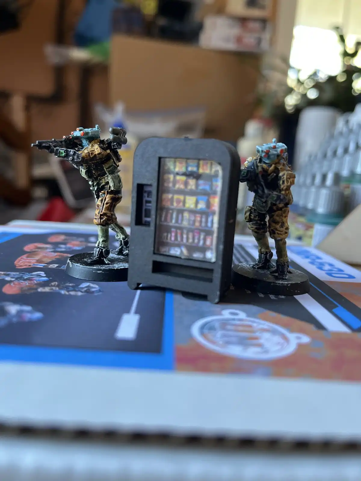 Two painted miniature sci-fi soldiers flank a tiny model vending machine filled with snack items. The soldier on the left…