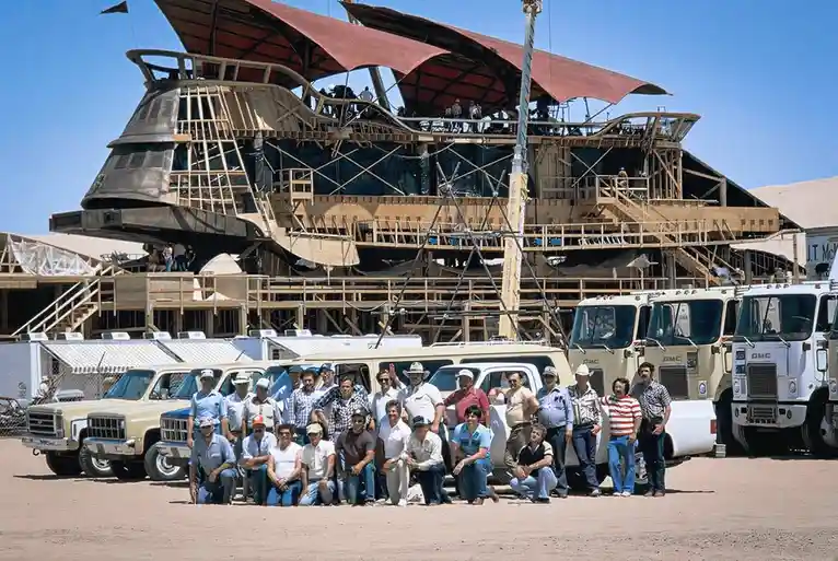 A group of people pose for a photo in front of a large, unfinished movie set resembling a spaceship or futuristic structure…