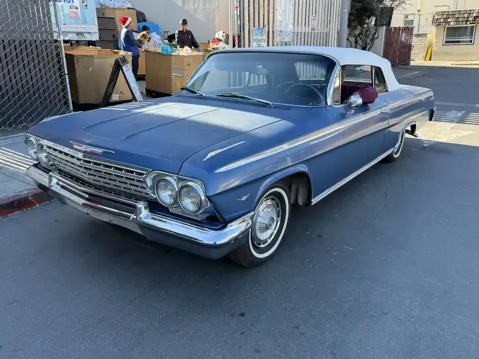 A blue 1960s Chevrolet convertible with a white roof is parked on a street, its driver's side window down. A person in a…