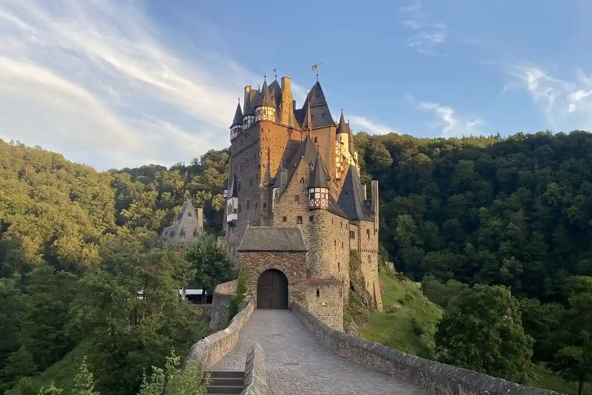 A majestic, multi-turreted stone castle with dark roofs and wooden-framed windows sits atop a lush green hillside, viewed…