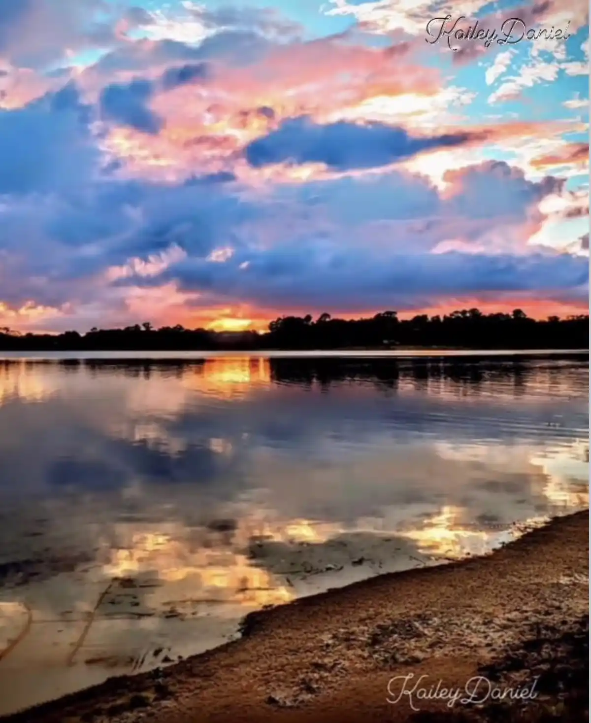 A serene sunset over a calm lake with vibrant pink, orange, and blue clouds reflected in the water, framed by a distant tree…