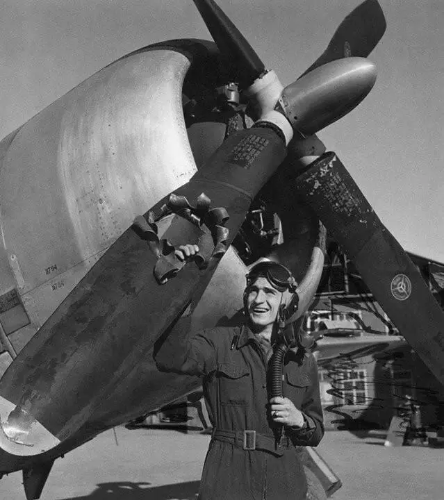 A black-and-white photo shows a smiling pilot in flight gear standing beside a large propeller aircraft, holding onto the…