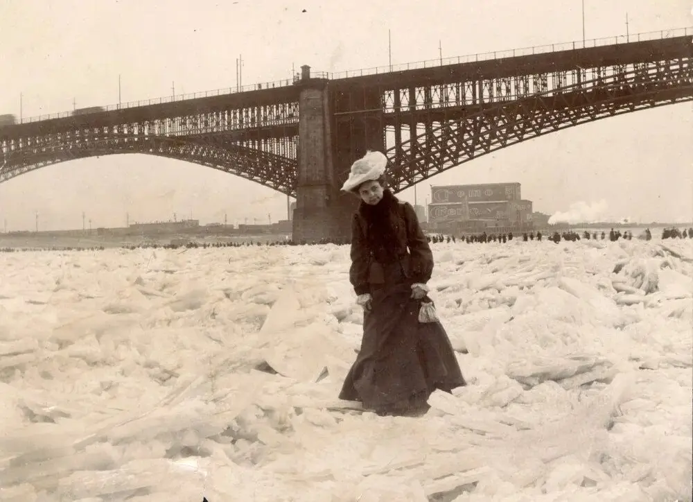 A vintage black-and-white photograph shows a woman in a long dark coat and white hat standing confidently on a field of…