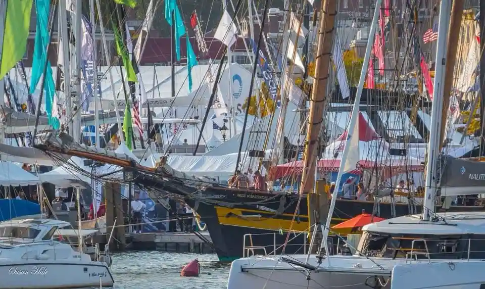 A crowded marina filled with sailboats and yachts, some with masts and rigging visible, people walking on docks, and tents…