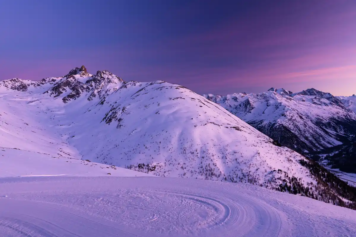 A breathtaking photo of snow-covered mountains under a purple and blue twilight sky, with ski tracks curving across the…