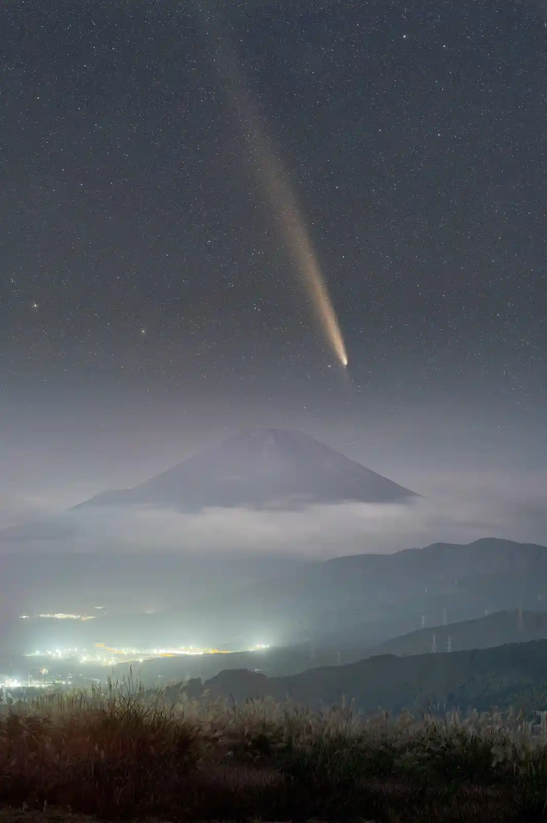 A night photograph shows Mount Fuji partially obscured by clouds, with a bright comet streaking across the starry sky above…