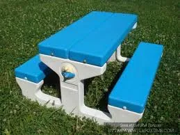 a bright blue plastic picnic table with white legs sits on green grass, featuring a small circular logo on the side of the…
