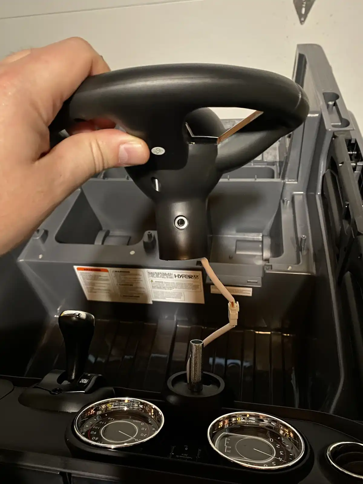 A hand grips a black steering wheel of a toy or miniature vehicle, which is visibly detached and hanging by a wire from its…