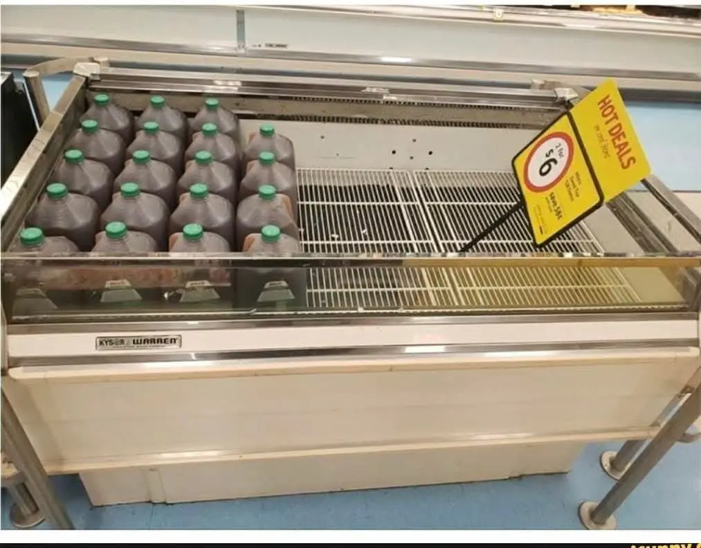 A supermarket freezer display contains only a few large plastic bottles of dark liquid with green caps, while the rest of…