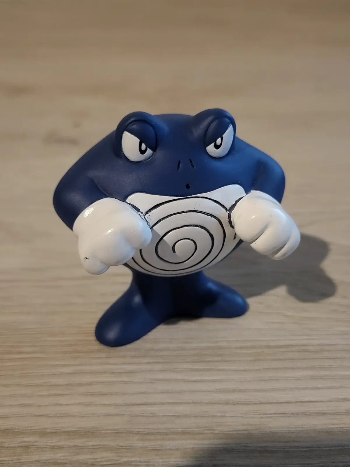 A blue Pokemon figurine, identified as Poliwrath, stands on a wooden surface with a stern, angry expression and clenched…