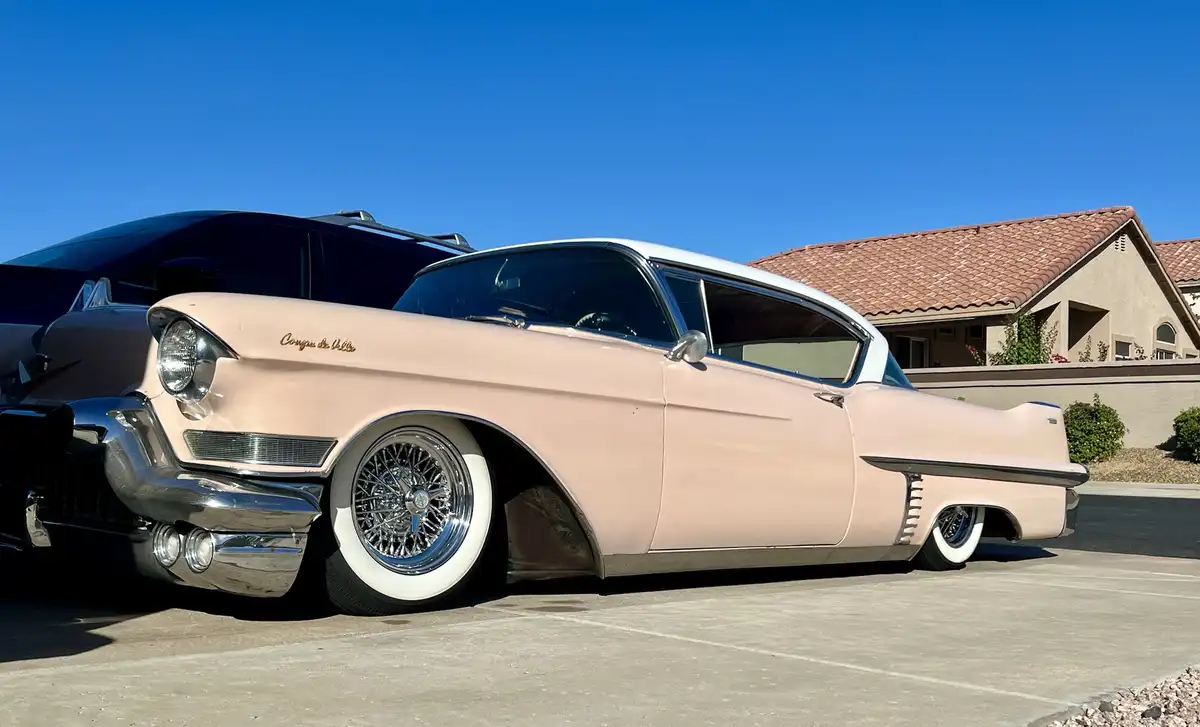 A light pink 1950s Cadillac Coupe de Ville with chrome details and whitewall tires is parked in a driveway under a clear…