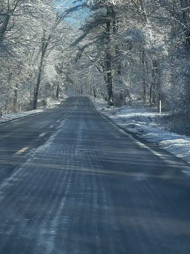 A straight, empty asphalt road stretches into the distance through a snowy forest, with trees heavily coated in white snow…