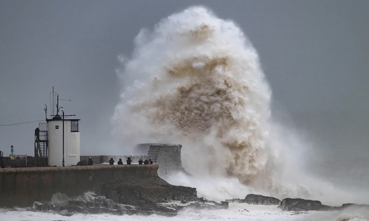 A massive, turbulent wave crashes against a stone sea wall, sending a huge plume of white and brown water skyward. Several…