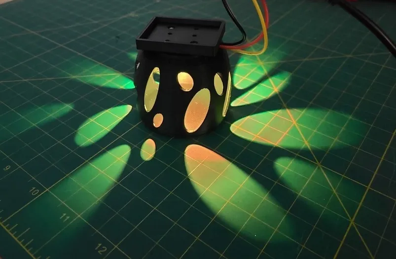 A black, cube-shaped object with cut-out oval holes sits on a green grid cutting mat, casting colorful, leaf-like light…