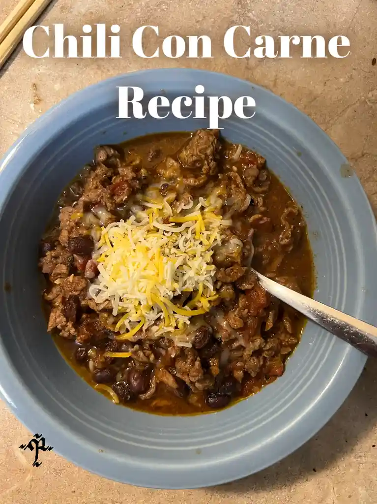 A top-down photo of a blue bowl filled with chili con carne topped with shredded cheese, with a spoon resting in the bowl…