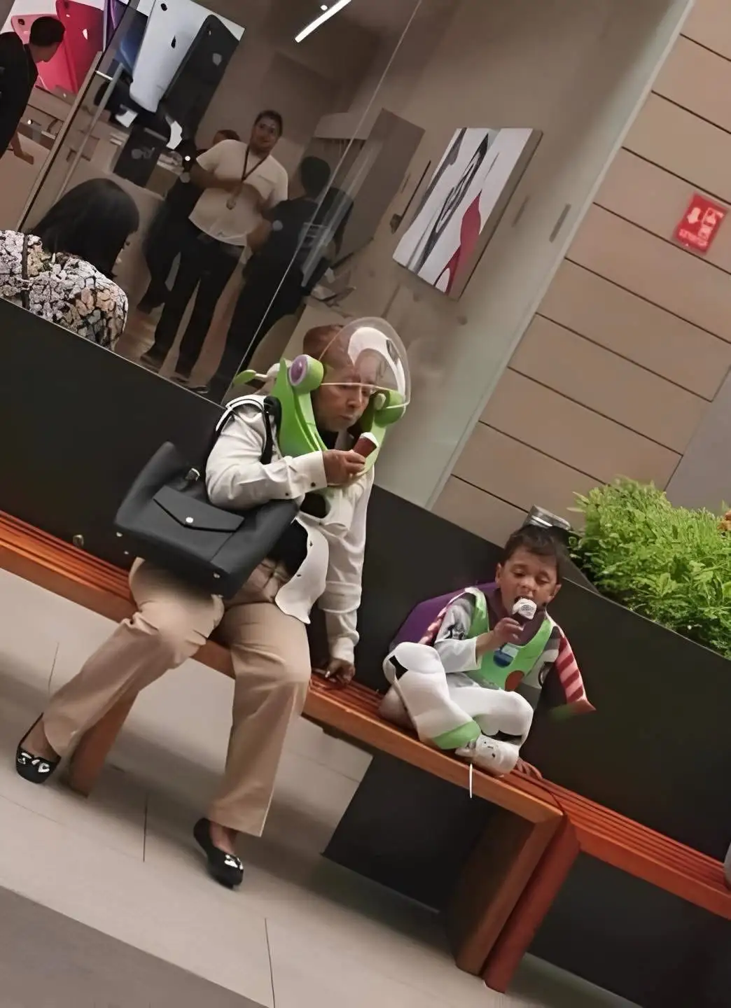 An older woman and a young boy are sitting on a bench, both wearing Buzz Lightyear costumes from Toy Story. The woman…