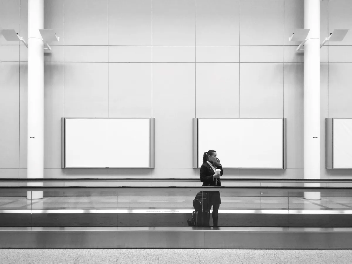 A black-and-white photo shows a woman in a suit standing on a baggage carousel in an airport, holding a coffee cup and…