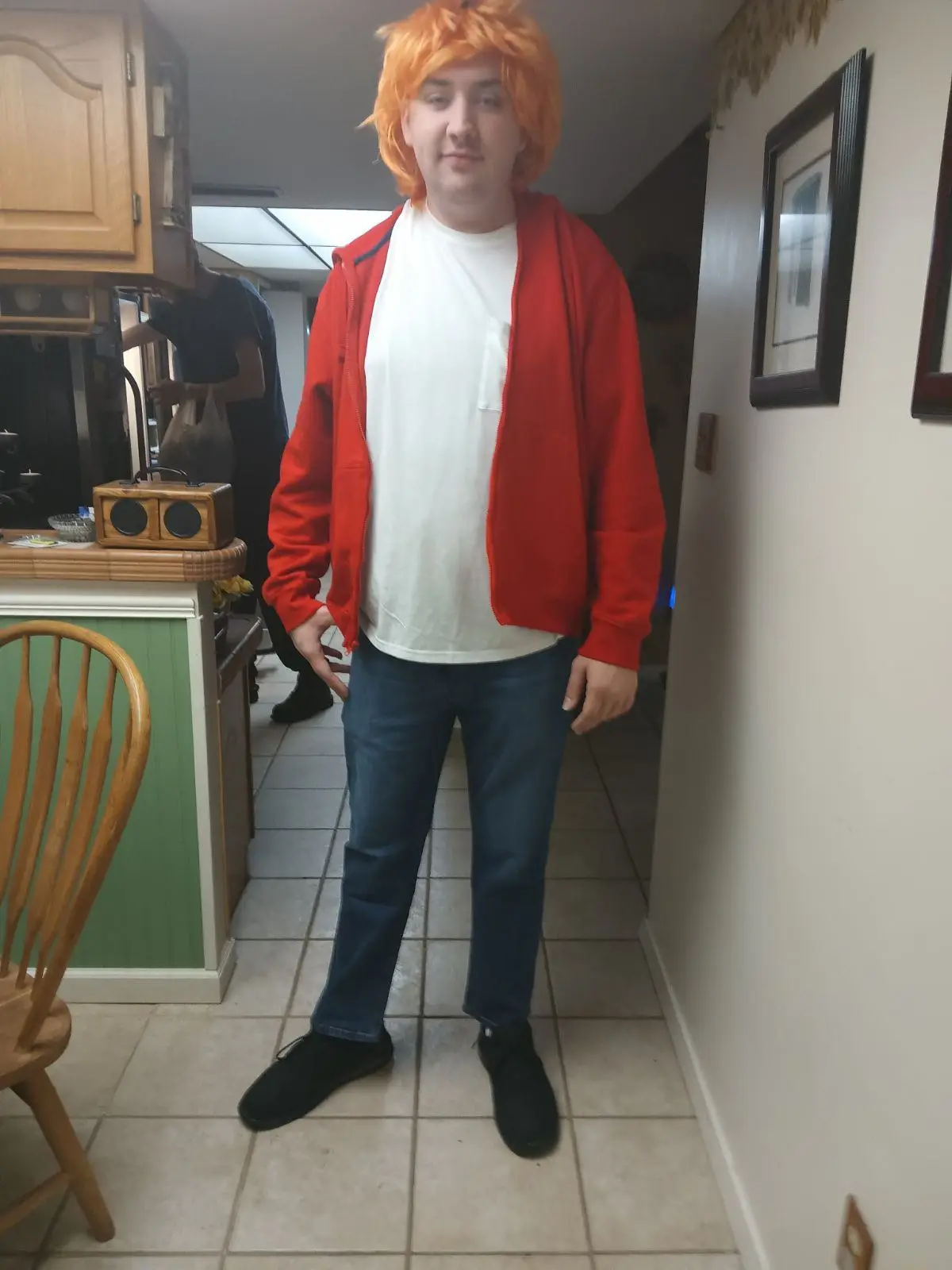 A person stands in a kitchen wearing an orange wig, a red zip-up hoodie over a white t-shirt, and blue jeans. They have a…