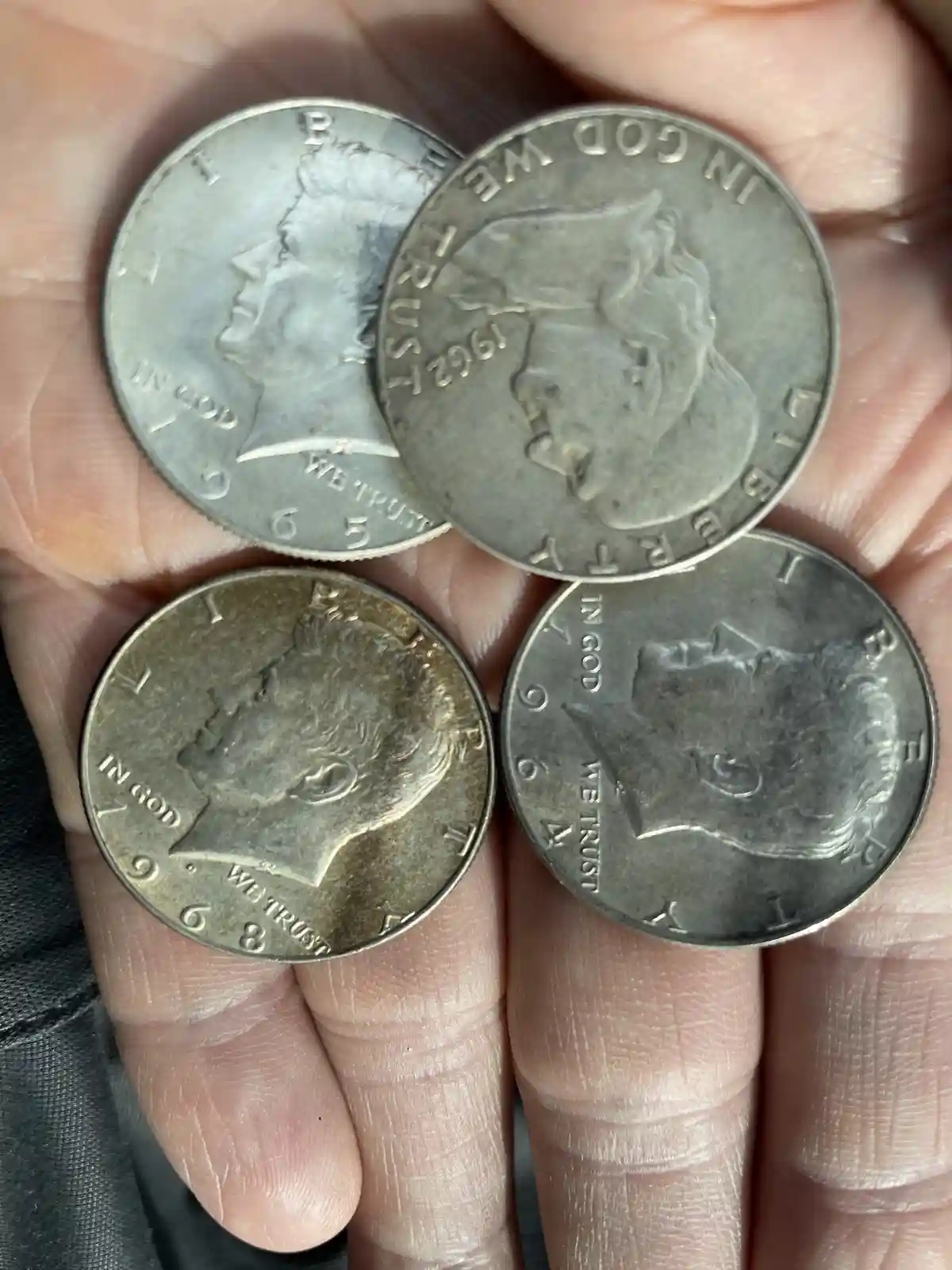 A hand holds four silver-colored U.S. coins, all showing the 'In God We Trust' motto and dated 1965, 1962, and 1968. The…
