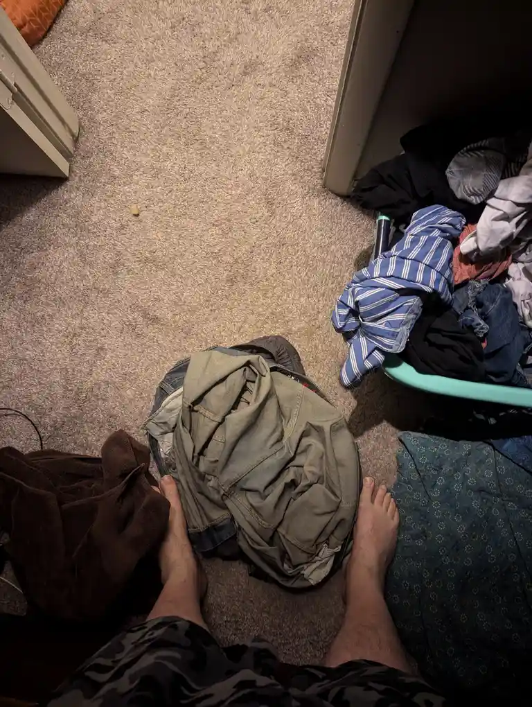 A first-person view shows bare feet on a carpeted floor surrounded by piles of unfolded laundry, including a large heap of…
