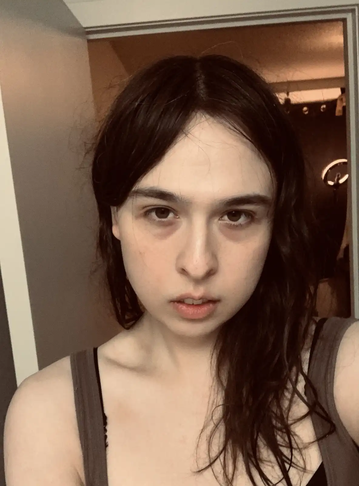 A person with dark, wet-looking hair and a tired expression stares directly at the camera, wearing a tank top. Behind them…