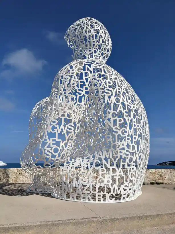 A large white sculpture of a seated figure, constructed from numerous cut-out letters and words, is shown against a blue sky…
