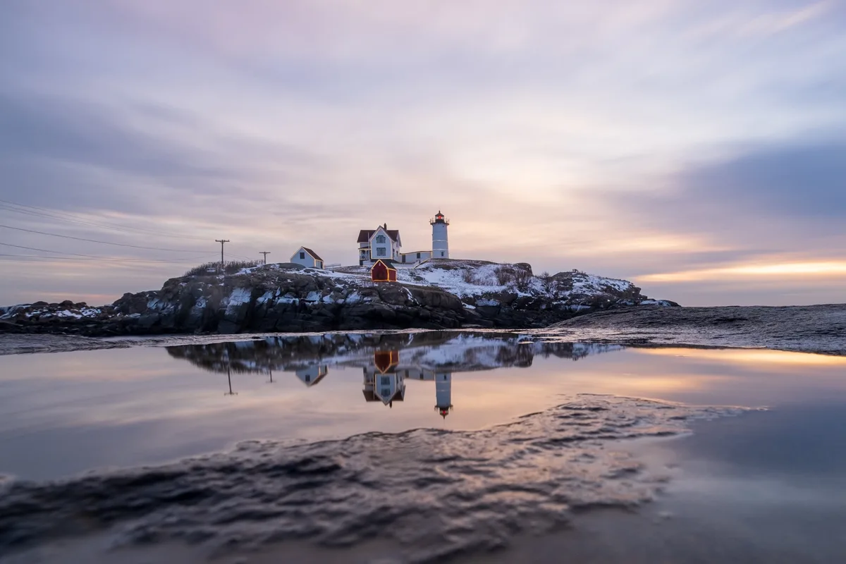 A serene winter landscape featuring a lighthouse and adjacent buildings on a snow-dusted rocky island, reflected perfectly…