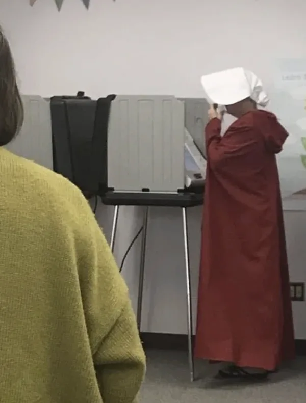 A person wearing a red robe and white bonnet, resembling a character from 'The Handmaid's Tale', stands at a voting booth…