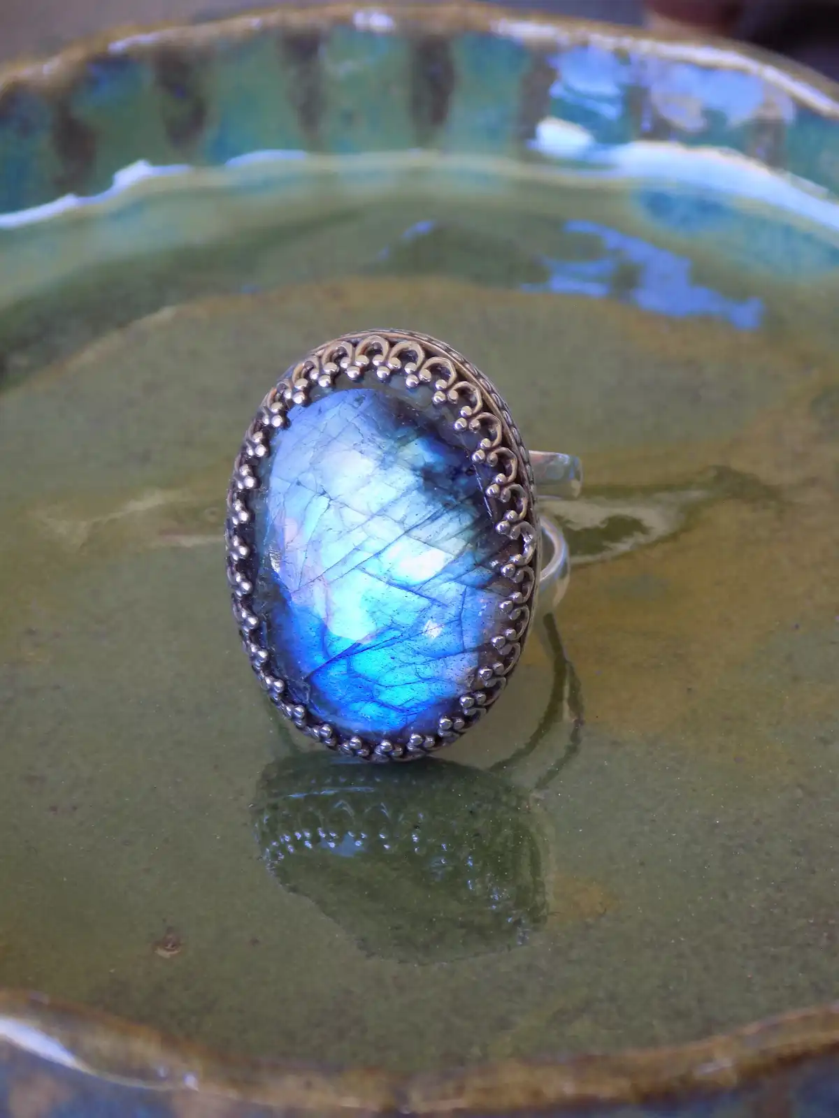 A close-up photo shows a silver ring with an oval, iridescent blue labradorite gemstone resting in a green ceramic bowl. The…