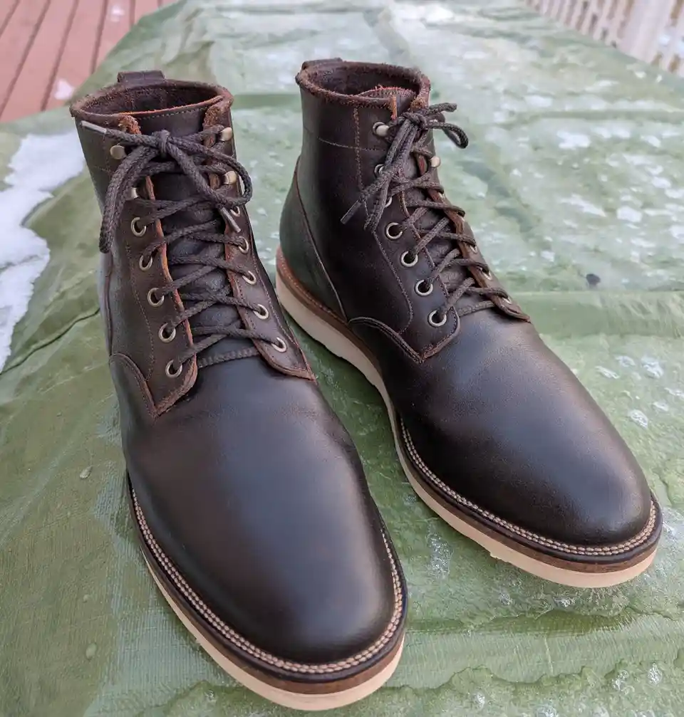 A pair of dark brown leather lace-up boots with tan soles sits on a green tarp with patches of melting snow. The overlaid…