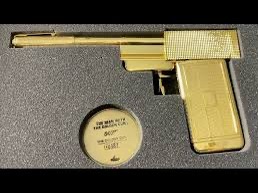 A golden-colored pistol rests in a gray foam-lined case next to a circular gold disc. The disc has text that reads 'THE MAN…