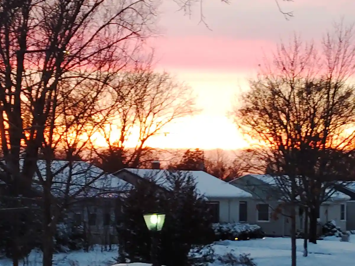 A snowy suburban neighborhood at sunset, with bare trees silhouetted against a vibrant orange and pink sky, houses covered…