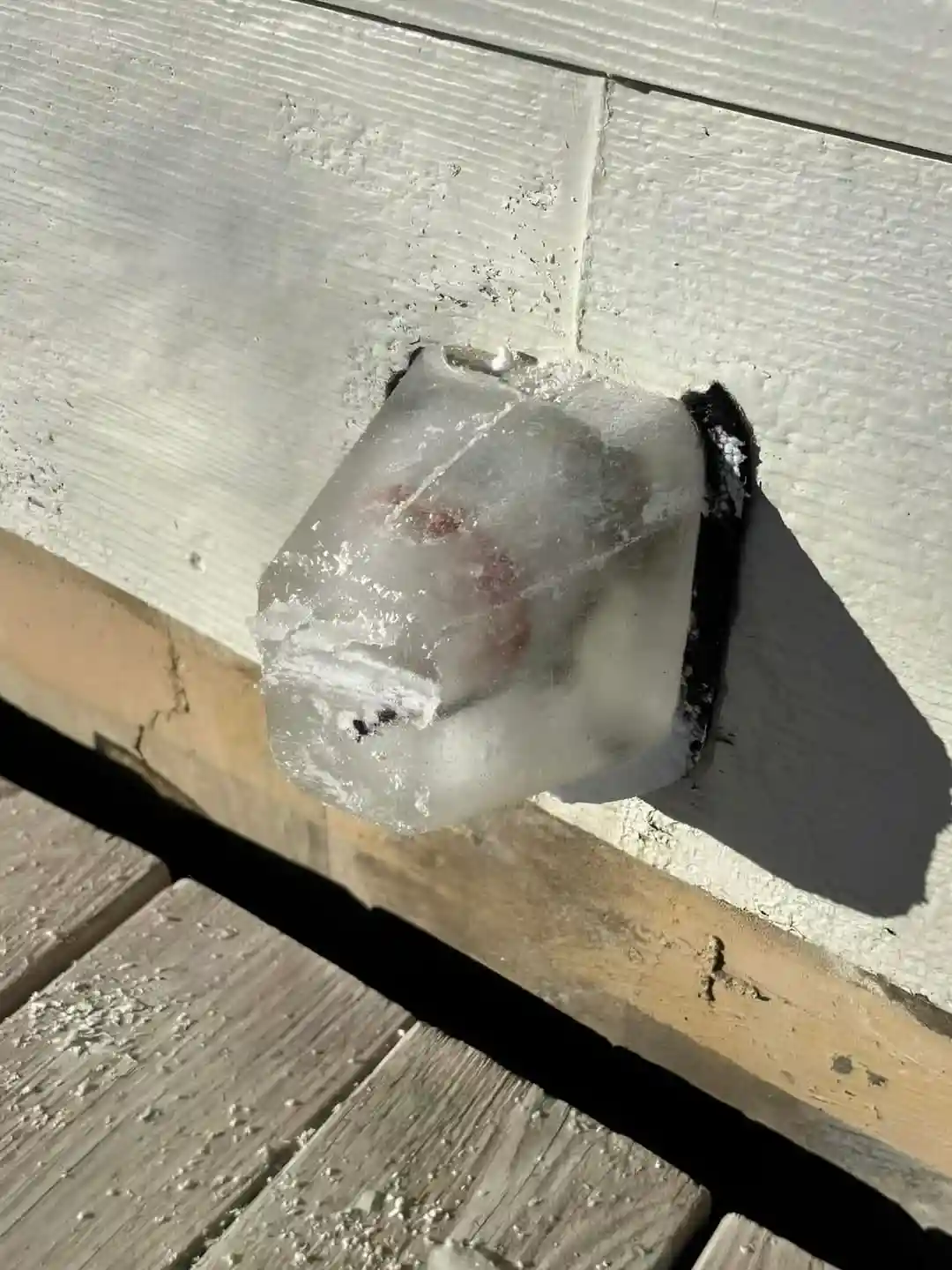 A large, clear ice cube is frozen in a hole in a white wooden wall, with a reddish-brown object visible inside. The ice cube…