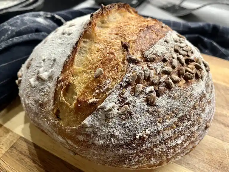 A close-up photo of a round, rustic sourdough loaf with a golden-brown crust, dusted with flour and topped with sunflower…