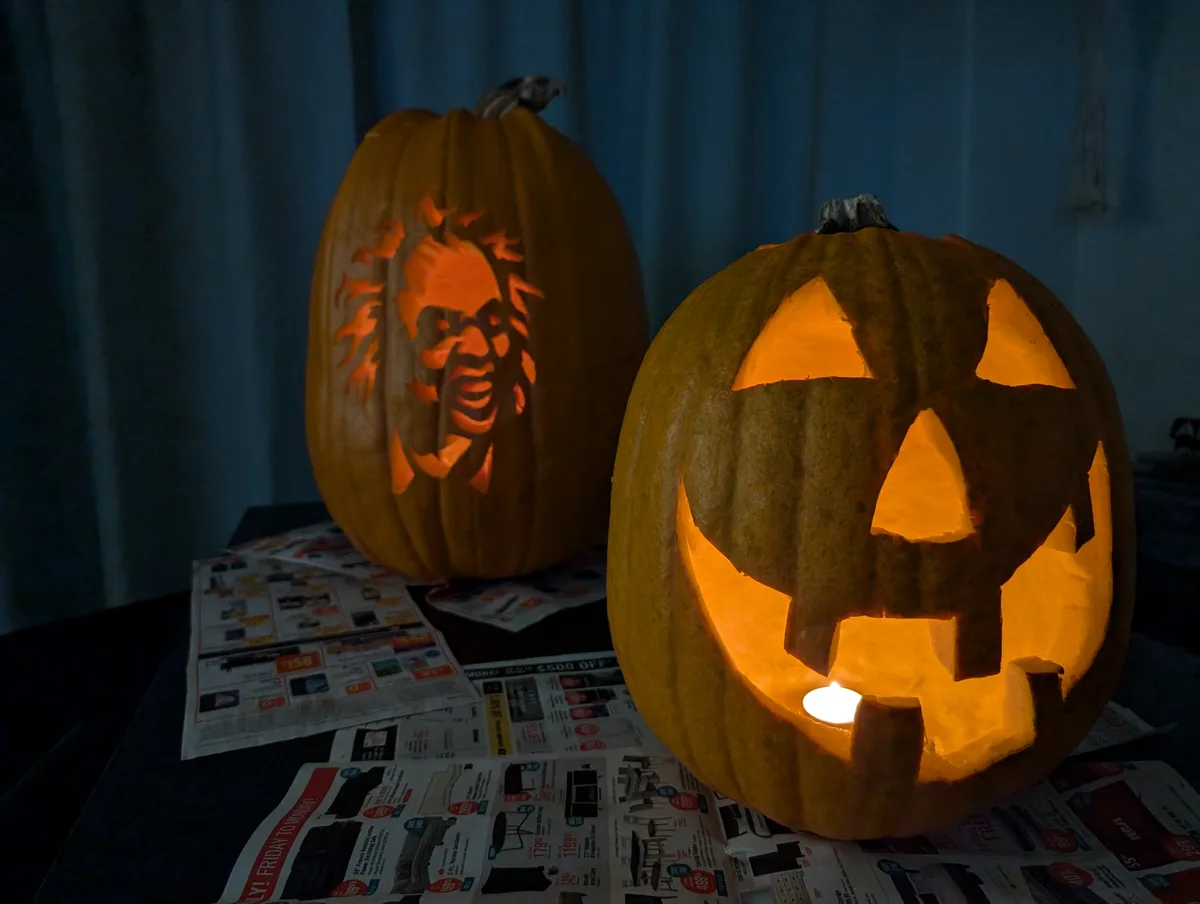 Two carved pumpkins sit on a table covered with newspaper ads in a dimly lit room. The pumpkin in the foreground has a…