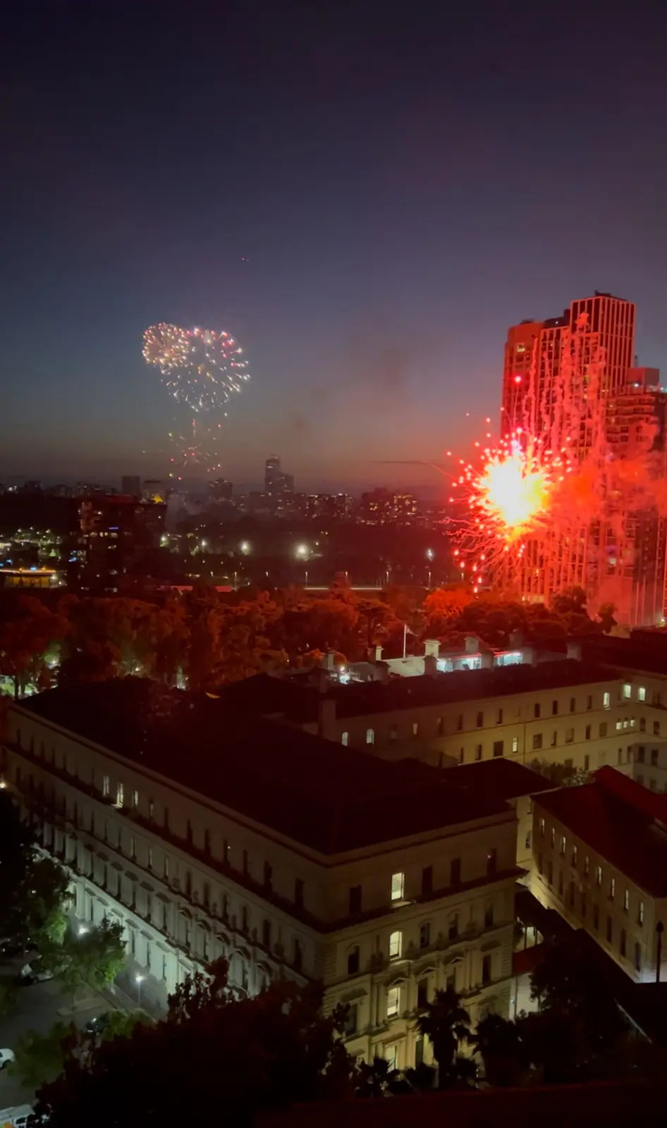 A nighttime cityscape photo shows two distinct firework displays: one colorful, heart-shaped burst on the left and a large…