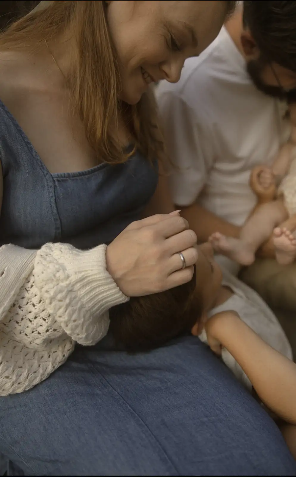 A woman with reddish-brown hair, wearing a denim top and white knit sleeves, smiles while gently touching the head of a…