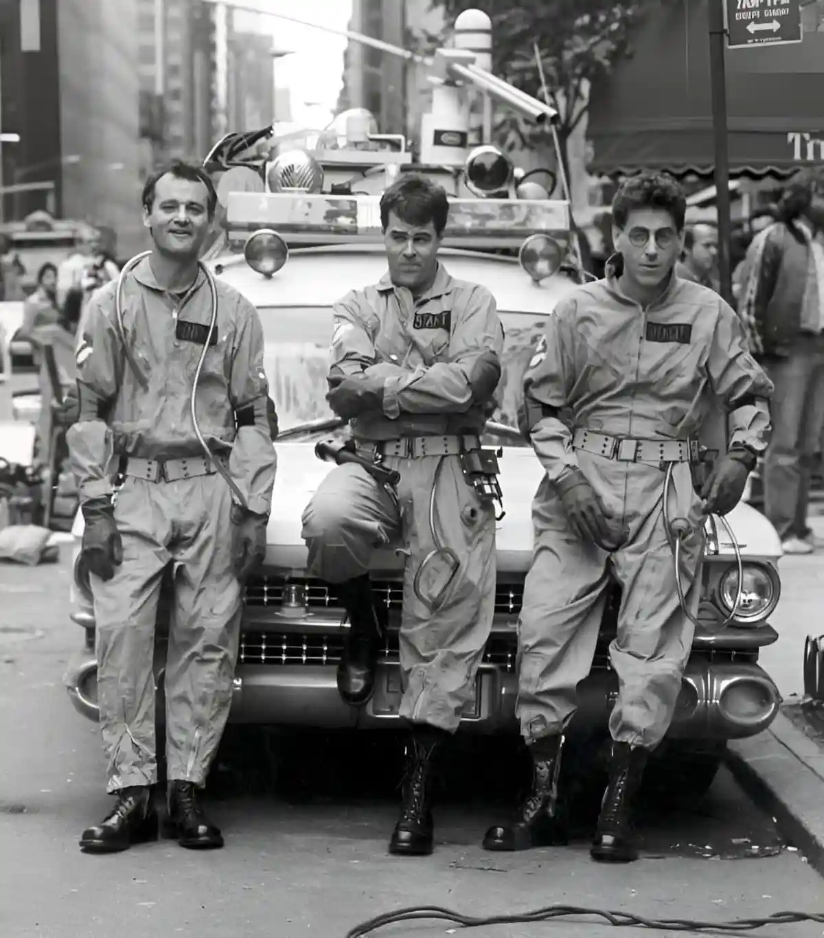 Three men in Ghostbusters uniforms stand confidently in front of their iconic Ecto-1 vehicle on a city street. The man on…